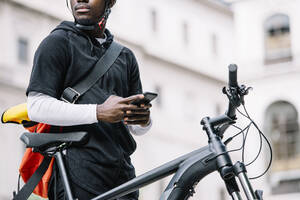 Stylischer junger Mann mit Fahrrad, Smartphone und Umhängetasche in der Stadt - CJMF00161
