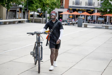 Stilvoller junger Mann mit Umhängetasche, der ein Fahrrad in der Stadt schiebt - CJMF00163