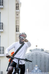 Stilvoller junger Mann mit Fahrrad und Umhängetasche in der Stadt, der mit dem Finger zeigt - CJMF00166