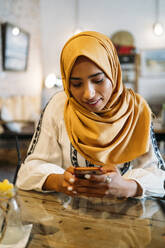 Junge muslimische Frau mit gelbem Hidschab in einem Café und mit Smartphone - MPPF00210