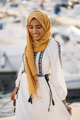 Junge muslimische Frau mit gelbem Hidschab beim Spaziergang am Hafen, lizenzfreies Stockfoto