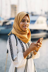Junge muslimische Frau mit gelbem Hidschab beim Spaziergang am Hafen - MPPF00218