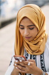 Junge muslimische Frau trägt gelben Hijab und benutzt ein Smartphone - MPPF00222