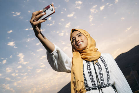 Junge muslimische Frau, die einen gelben Hidschab trägt und ein Smartphone benutzt und ein Selfie am Abend macht, lizenzfreies Stockfoto