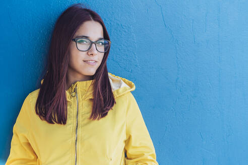 Lächelndes Teenager-Mädchen mit gelber Regenjacke vor einer blauen Wand - DLTSF00270