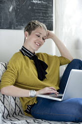 Portrait of smiling woman sitting on couch looking at laptop - FLLF00312