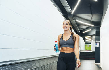 Lächelnde Frau in guter Form, die während des Trainings mit einer Wasserflasche durch einen Korridor läuft - JCMF00256