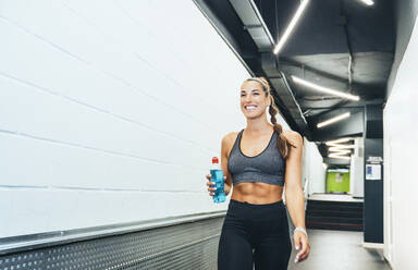 Smiling woman in good shape walking through corridor during workout with water bottle - JCMF00256