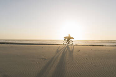 Surfer auf einem Fahrrad während des Sonnenuntergangs am Strand, Costa Nova, Portugal - AHSF01043