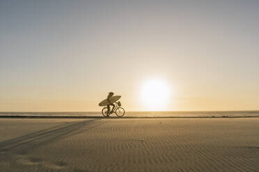 Surfer auf einem Fahrrad während des Sonnenuntergangs am Strand, Costa Nova, Portugal - AHSF01044