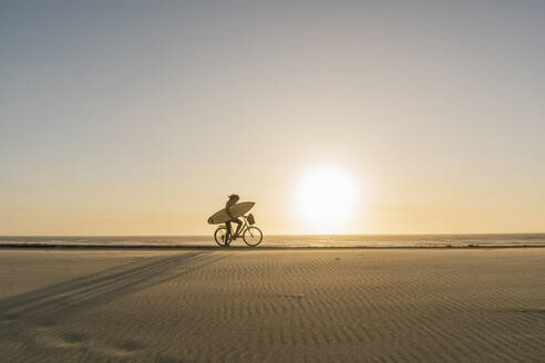 Surfer riding a bicycle during the sunset in the beach, Costa Nova, Portugal - AHSF01044