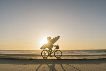 Surfer auf einem Fahrrad während des Sonnenuntergangs am Strand, Costa Nova, Portugal - AHSF01045