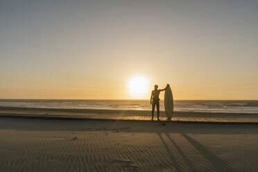 Surfer standing with the surfboard facing the sunset at the beach - AHSF01047