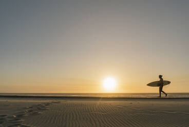 Surfer, der während des Sonnenuntergangs am Strand mit dem Surfbrett spazieren geht - AHSF01049