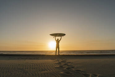 Surfer holding up surfboard on the beach, looking at sunset - AHSF01050