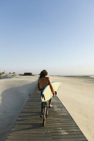 Surfer, der bei Sonnenuntergang Fahrrad fährt und ein Surfbrett hält, lizenzfreies Stockfoto