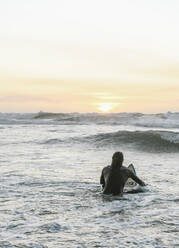 Rückansicht eines Surfers mit Surfbrett, der während des Sonnenuntergangs auf einem Surfbrett im Meer sitzt - AHSF01064