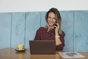 Woman talking on the phone in a cafe and looking at laptop - MOMF00787