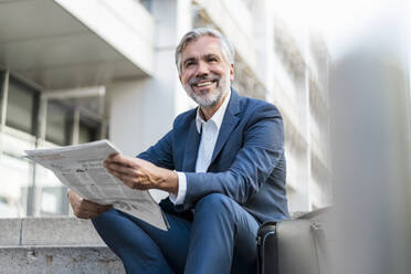 Lächelnder reifer Geschäftsmann, der mit einer Zeitung auf einer Treppe in der Stadt sitzt - DIGF08531