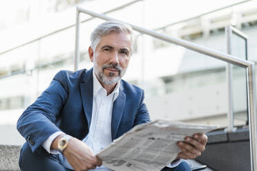 Portrait of mature businessman sitting on stairs in the city with newspaper - DIGF08535