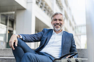 Portrait of smiling mature businessman sitting on stairs in the city - DIGF08537