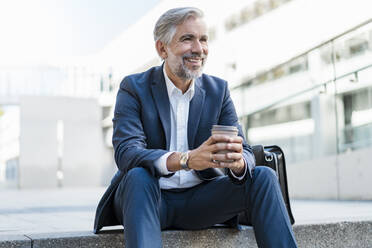 Smiling mature businessman sitting on stairs in the city holding takeaway coffee - DIGF08538