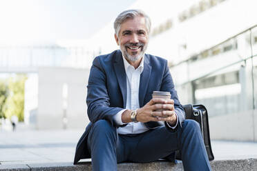 Portrait of smiling mature businessman sitting on stairs in the city holding takeaway coffee - DIGF08539