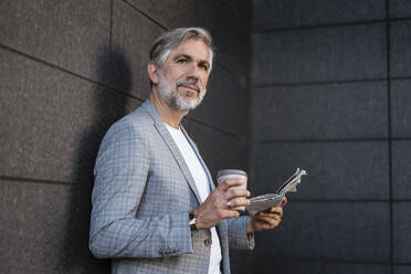 Fashionable mature businessman leaning at a wall with newspaper and takeaway coffee - DIGF08579