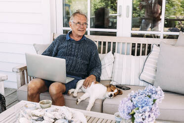 Mann mit Laptop, sitzt neben Hund auf Sofa - JOHF04463