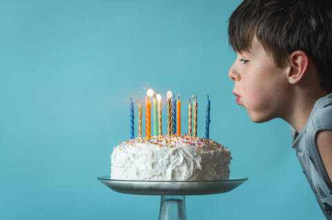 Junge bläst Kerzen auf einer Geburtstagstorte vor blauem Hintergrund aus., lizenzfreies Stockfoto