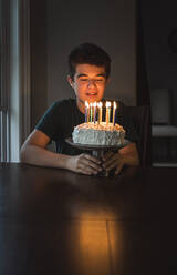 Teenager, der die Kerzen auf seinem Geburtstagskuchen in einem dunklen Raum betrachtet. - CAVF65891