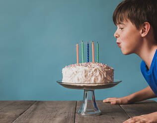 Junge bläst Kerzen auf einer Geburtstagstorte vor blauem Hintergrund aus. - CAVF65893