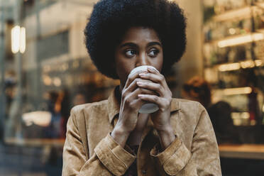 Junge Frau mit Afro-Haar, die vor einem Café ein heißes Getränk zu sich nimmt - CUF52566