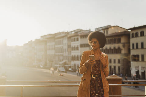 Junge Frau mit Afro-Haar, die ein heißes Getränk zu sich nimmt und ein Smartphone in der Stadt benutzt, Florenz, Toskana, Italien, lizenzfreies Stockfoto