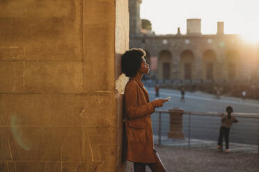 Junge Frau mit Afro-Haar, die ein Smartphone benutzt, an eine Steinmauer gelehnt, Florenz, Toskana, Italien - CUF52571