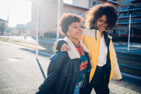 Zwei coole junge Freundinnen schlendern auf einem sonnenbeschienenen Bürgersteig, lizenzfreies Stockfoto