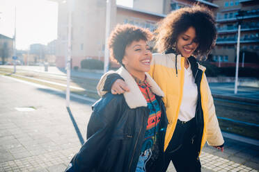 Zwei coole junge Freundinnen schlendern auf einem sonnenbeschienenen Bürgersteig - CUF52778