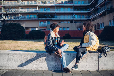 Zwei coole junge Freundinnen sitzen auf einer Stadtmauer und unterhalten sich - CUF52780
