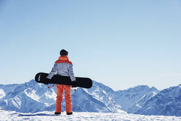 Jugendlicher Snowboarder, der von einem schneebedeckten Berggipfel auf die Landschaft blickt, Rückansicht, Alpe-d'Huez, Rhone-Alpes, Frankreich - CUF52820