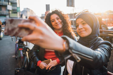 Young woman in hijab with friend taking smartphone selfie in city - CUF52823