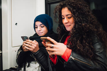 Young woman in hijab and friend sitting on subway train looking at smartphones - CUF52824