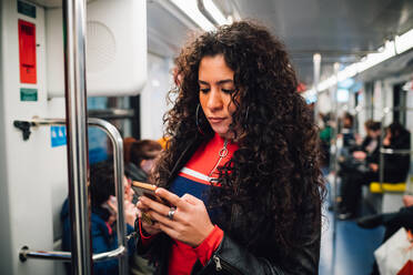 Mid adult woman with long curly hair looking at smartphone on city subway train - CUF52827