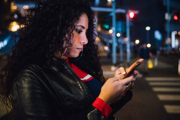 Mid adult woman with long curly hair looking at smartphone on city street at night - CUF52835