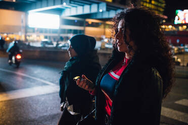 Junge Frau im Hidschab und Freundin auf dem Fußgängerüberweg in der Stadt bei Nacht - CUF52836