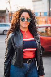 Mid adult woman with long curly hair wearing shades on city sidewalk, portrait - CUF52840