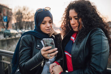 Junge Frau im Hidschab und beste Freundin schauen auf einer Kanalbrücke in der Stadt auf ihr Smartphone - CUF52842