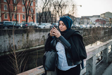 Junge Frau im Hidschab schaut am Kanal in der Stadt auf ihr Smartphone - CUF52844