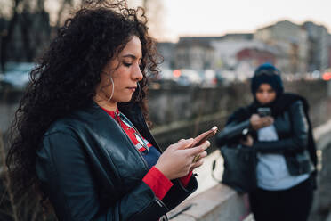 Mittlere erwachsene Frau mit langen lockigen Haaren schaut auf ihr Smartphone am Stadtkanal - CUF52845