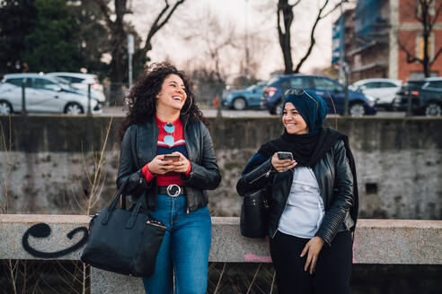 Junge Frau im Hidschab und ihre Freundin benutzen Smartphones und lachen am Stadtkanal - CUF52846