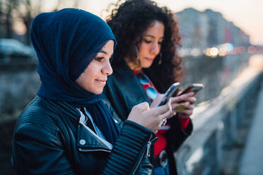 Junge Frau im Hidschab mit bester Freundin schaut auf Smartphones am Stadtkanal - CUF52852